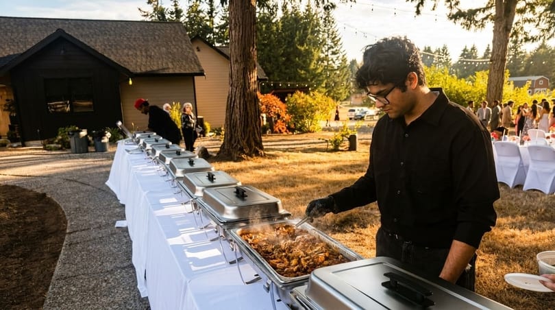 A caterer serves steaming food from a buffet line at an outdoor event with guests in the background.